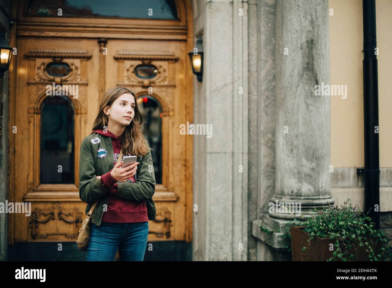 Nachdenkliches Mädchen hält Handy, während sie gegen Gebäude steht Stockfoto