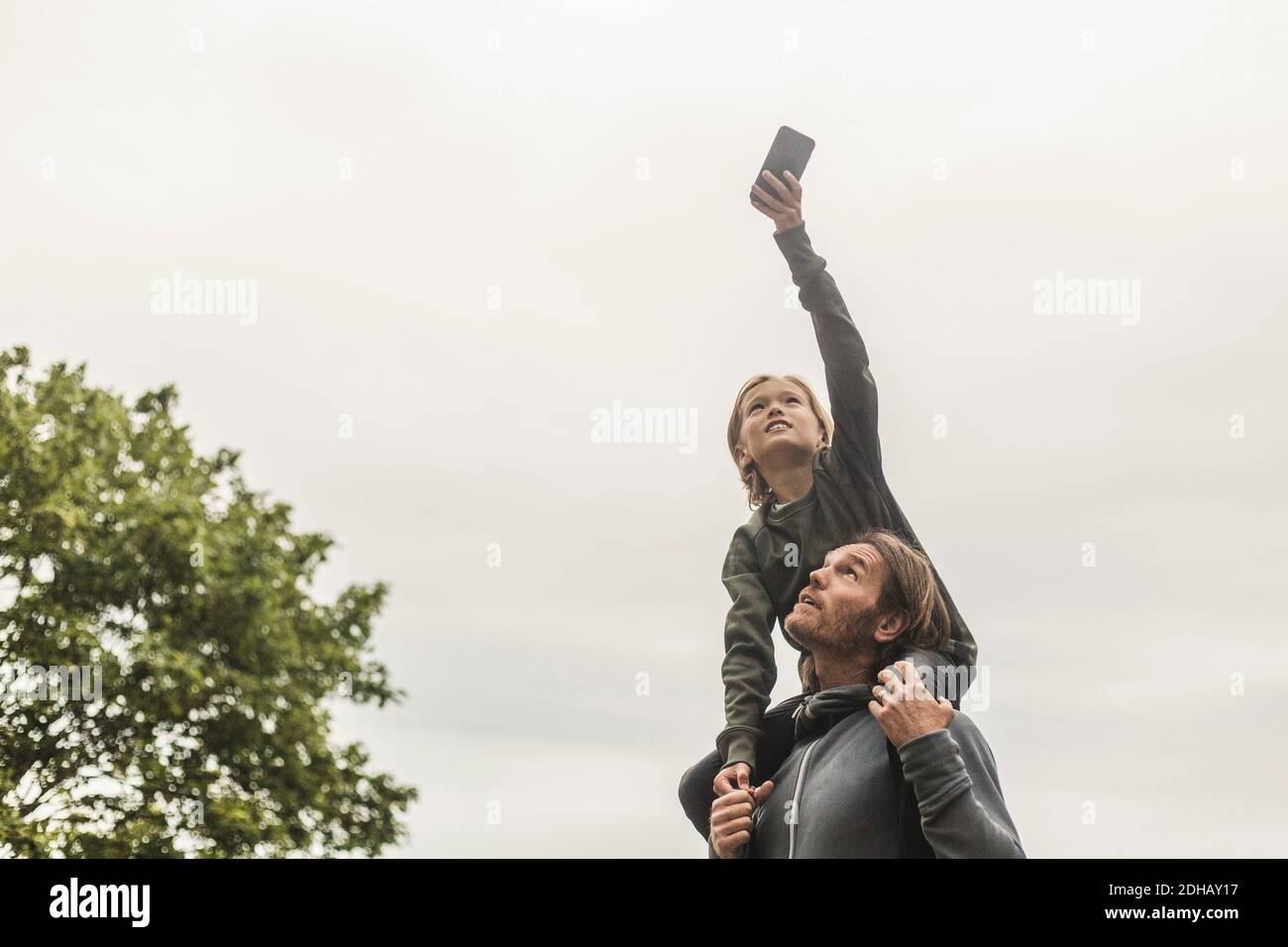 Tochter fotografiert durch Smartphone, während sie auf den Schultern des Vaters sitzt Gegen klaren Himmel Stockfoto
