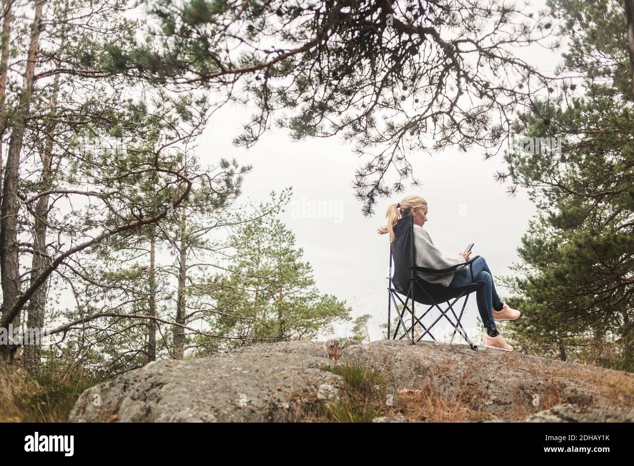Volle Länge der Frau mit Handy, während sie sitzt auf Campingstuhl über Felsformation gegen Bäume Stockfoto