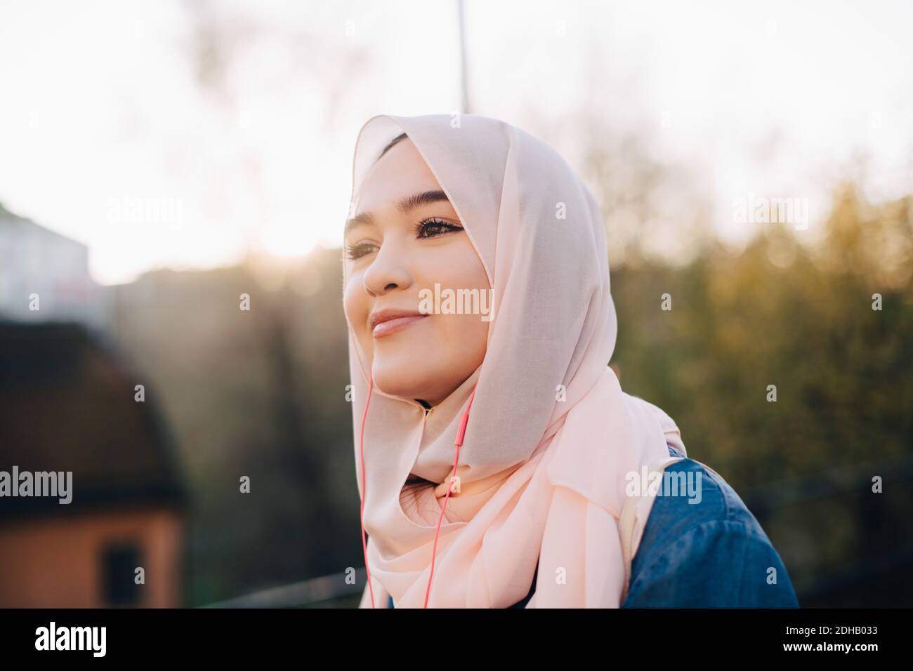 Lächelnde junge muslimische Frau, die Musik über in-Ear-Kopfhörer gegen Der Himmel ist klar Stockfoto