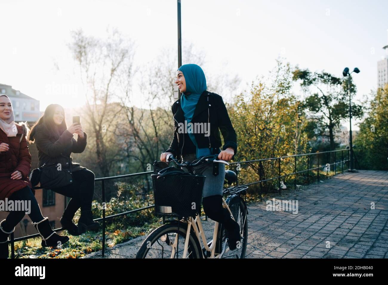 Lächelnde junge Frau, die auf dem Fußweg durch weibliche Freunde sitzt Auf Geländer in der Stadt Stockfoto