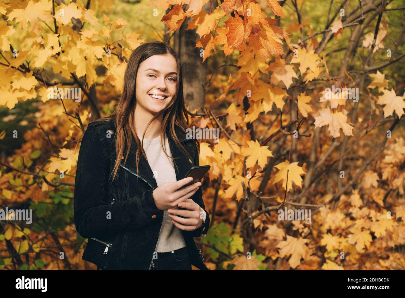 Portrait von lächelndem Teenager-Mädchen mit Handy, das gegen steht Bäume im Herbst Stockfoto