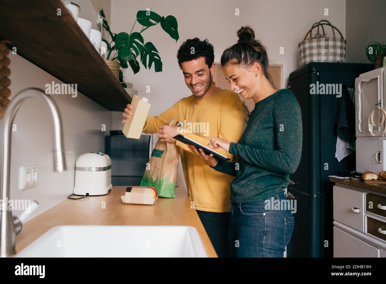 Lächelnder Mann, der Lebensmittel aus der Tasche herausgenommen hat, während Freundin digital zeigt Tablet an der Küchentheke Stockfoto