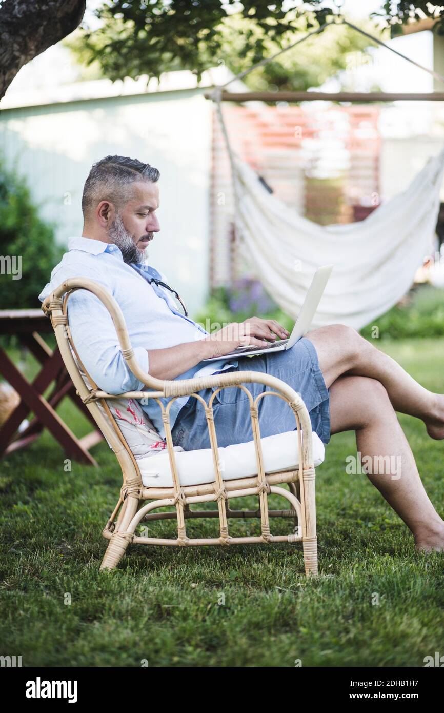 Seitenansicht eines reifen Mannes, der während des Sitzens am Laptop arbeitet Auf dem Stuhl im Hinterhof Stockfoto