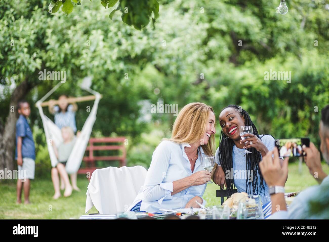 Mann mit Handy fotografiert lächelnde Frauen, die Wein trinken Gartenparty am Wochenende Stockfoto