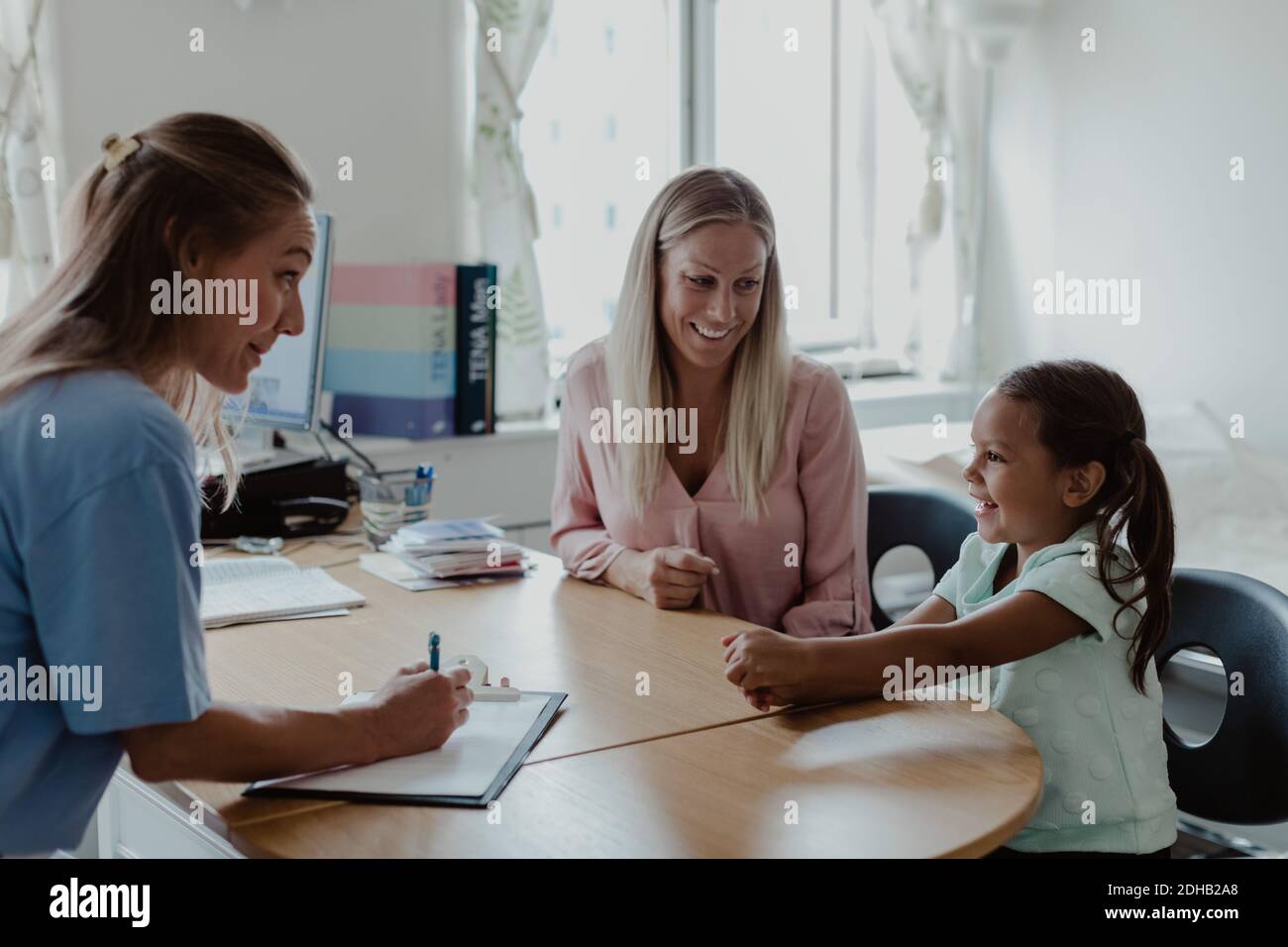 Kinderärztin und Mutter schauen lächelndes Mädchen an Schreibtisch im Krankenhaus Stockfoto