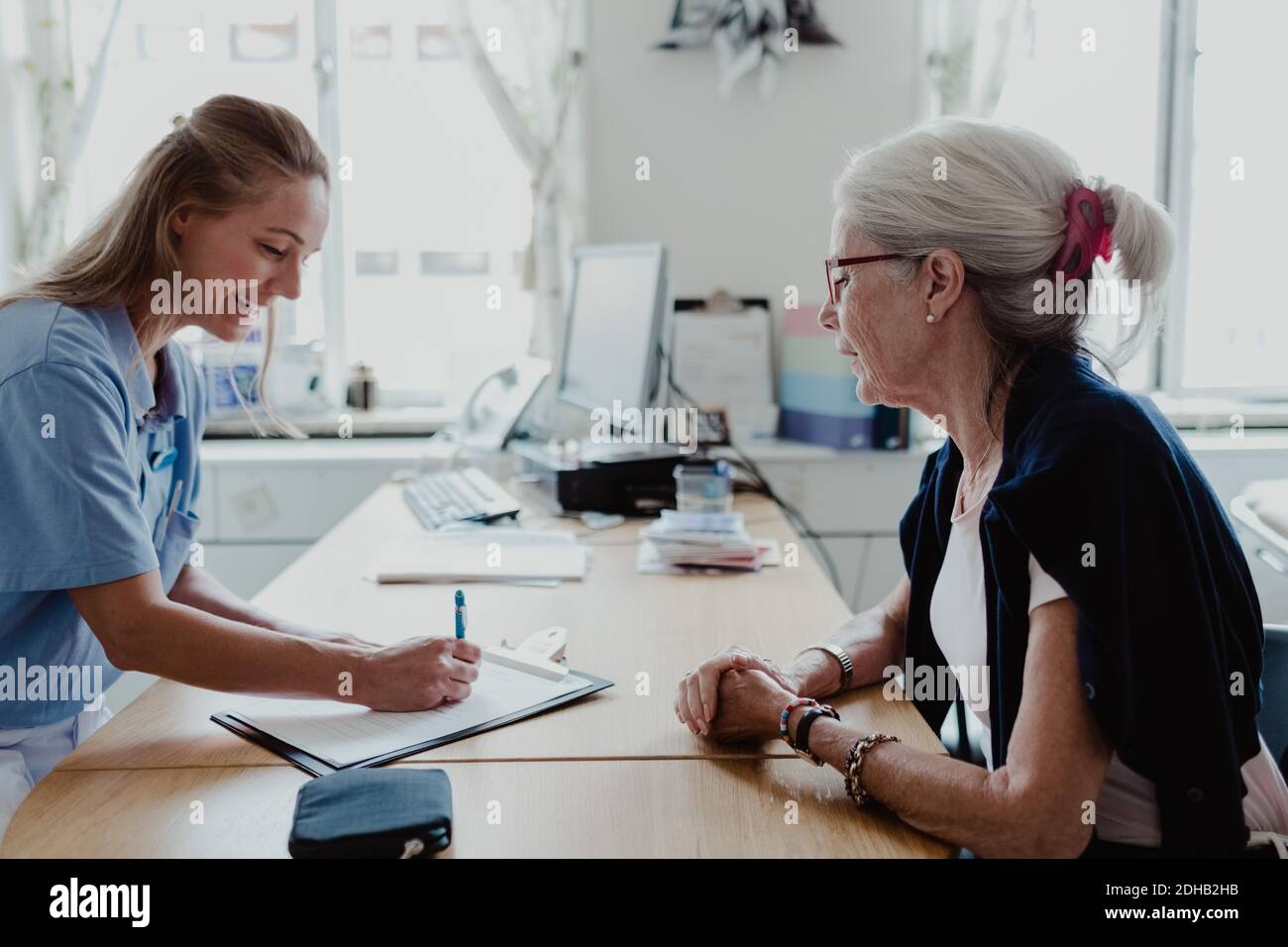 Lächelnder Arzt schreibt ein Rezept, während eine ältere Frau am Schreibtisch sitzt Im Krankenhaus Stockfoto