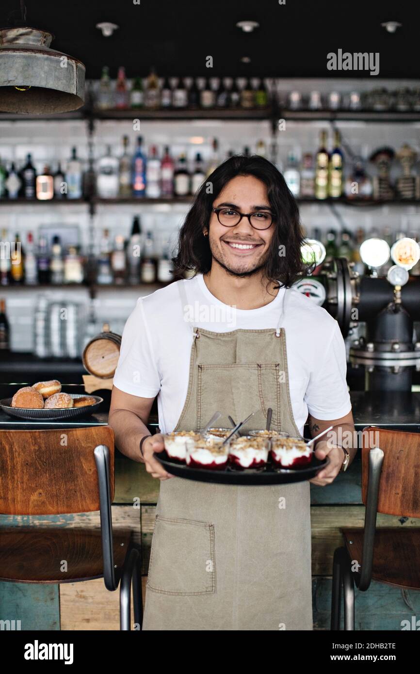 Porträt eines lächelnden Kellners, der während des Stehens Dessert im Tablett hält Gegen den Tresen im Restaurant Stockfoto