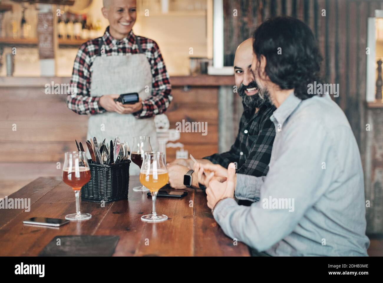 Barkeeper mit Kreditkartenleser im Gespräch mit männlichen Kunden Am Tisch in der Bar Stockfoto