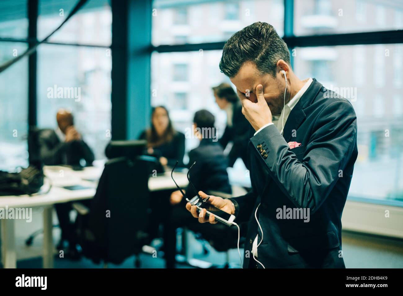 Gestresster Geschäftsmann mit Telefon über Kopfhörer, während Reiben Augen in Büro Stockfoto