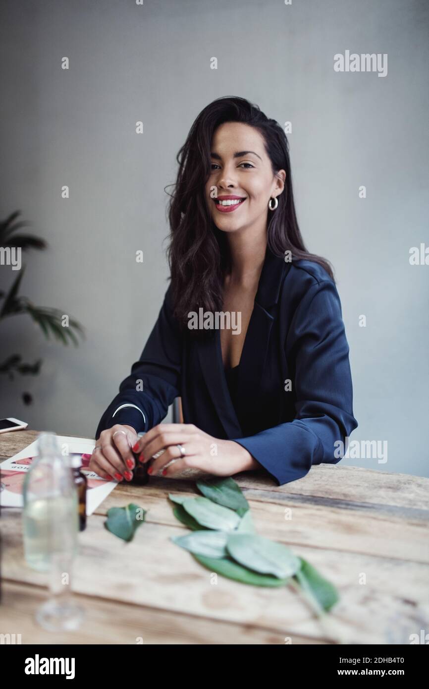 Porträt der lächelnden mittleren erwachsenen Unternehmerin am Tisch sitzen Gegen Wand in Parfümwerkstatt Stockfoto
