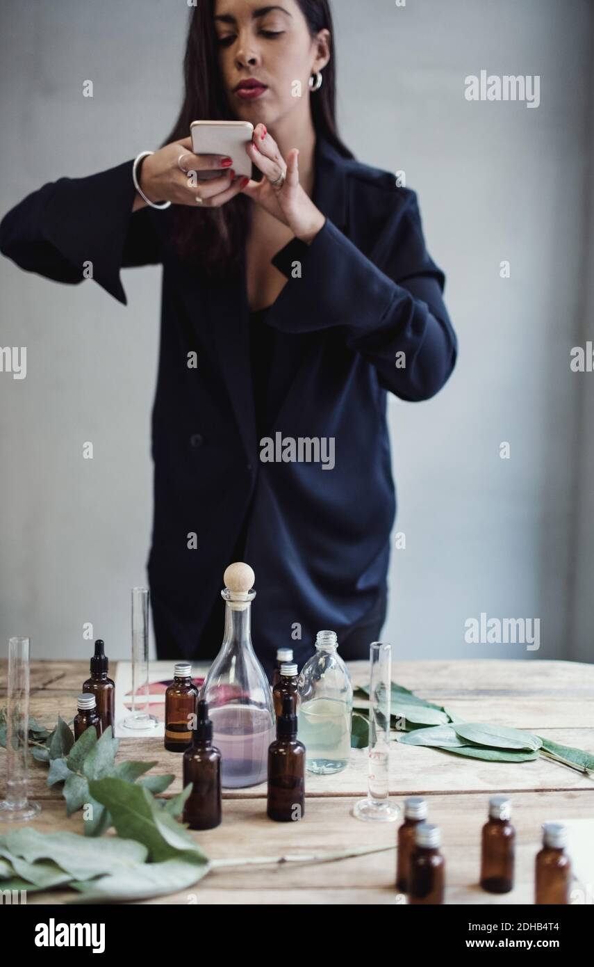 Mittlere Erwachsene weibliche Unternehmerin fotografiert Parfüm-Flaschen mit Smartphone Auf dem Tisch gegen die Wand in der Werkstatt Stockfoto