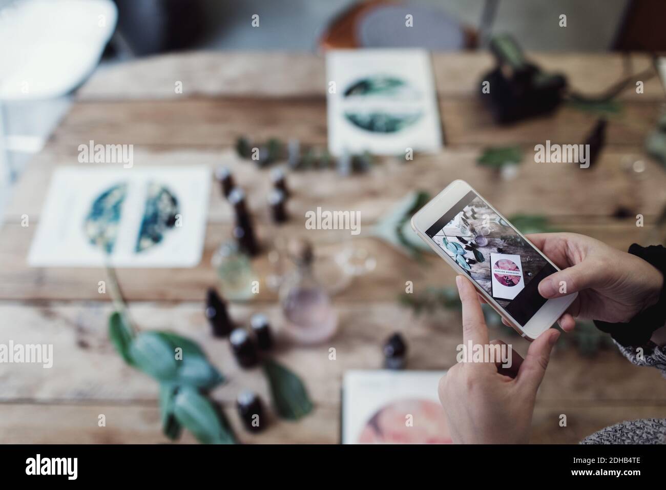 Beschnittene Hände der mittleren erwachsenen weiblichen Besitzerin, die Broschüre und fotografiert Parfümflaschen auf dem Tisch in der Werkstatt Stockfoto