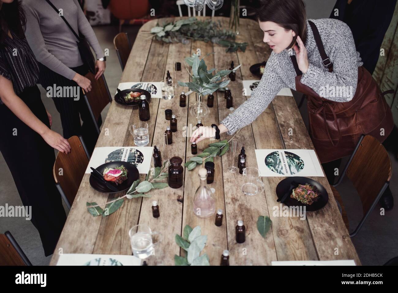 High-Angle-Ansicht der mittleren erwachsenen weiblichen Besitzerin hält Parfüm Flasche auf dem Tisch mit Kollegen, die in der Werkstatt stehen Stockfoto