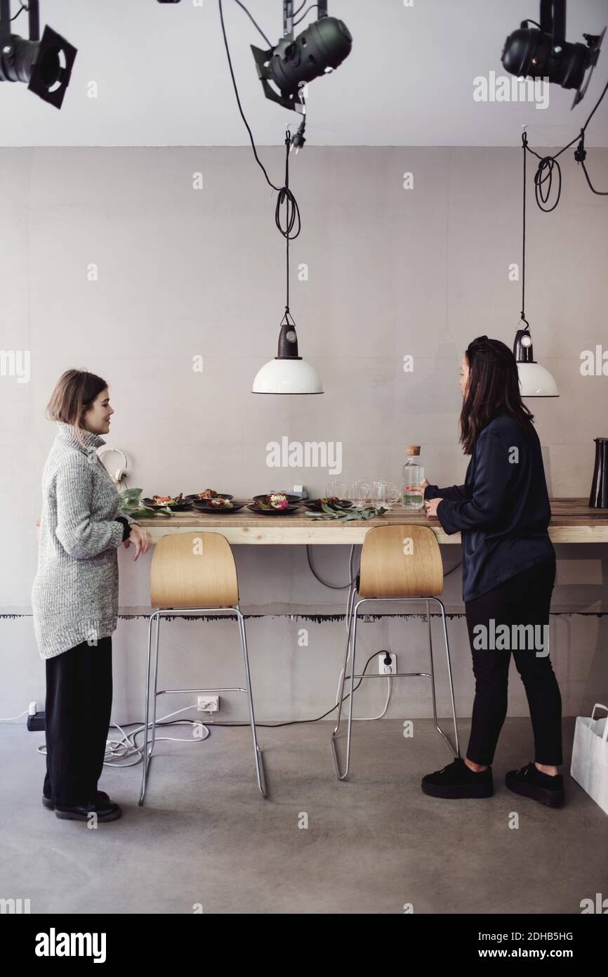 Volle Länge der mittleren erwachsenen weiblichen Kollegen neben dem Essen stehen Auf dem Tisch gegen die Wand in der Werkstatt Stockfoto