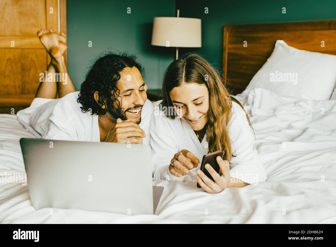 Ein Paar im Bademantel mit Laptop, das das Telefon benutzt, während man darauf liegt Bett im Hotelzimmer Stockfoto