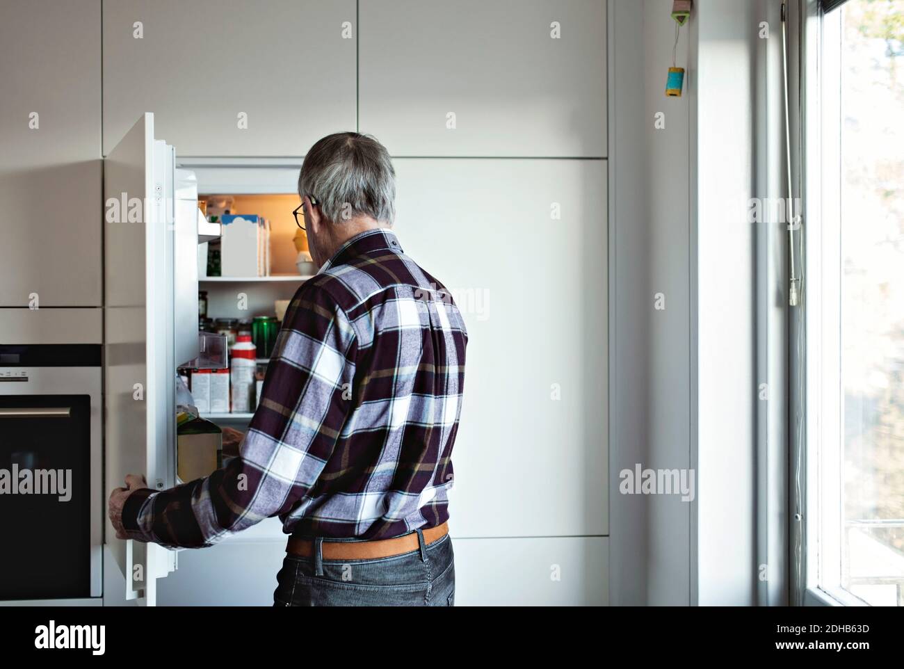 Rückansicht eines älteren Mannes im Ruhestand, der am offenen Kühlschrank stand Tür in der Küche zu Hause Stockfoto