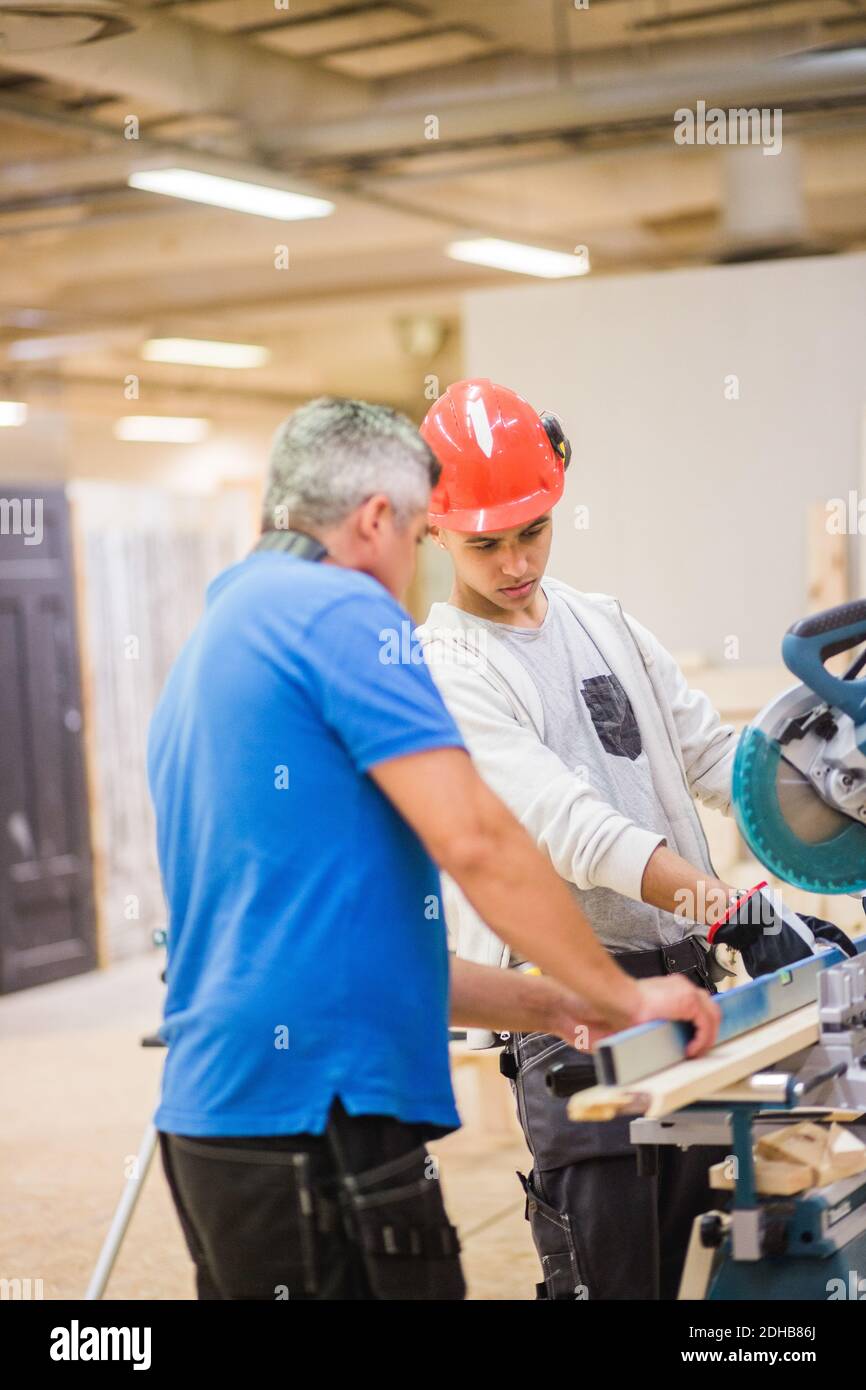 Männlicher Ausbilder, der den jungen Auszubildenden beim Schneiden der Planke mit elektrischer Säge hilft In der Werkstatt Stockfoto