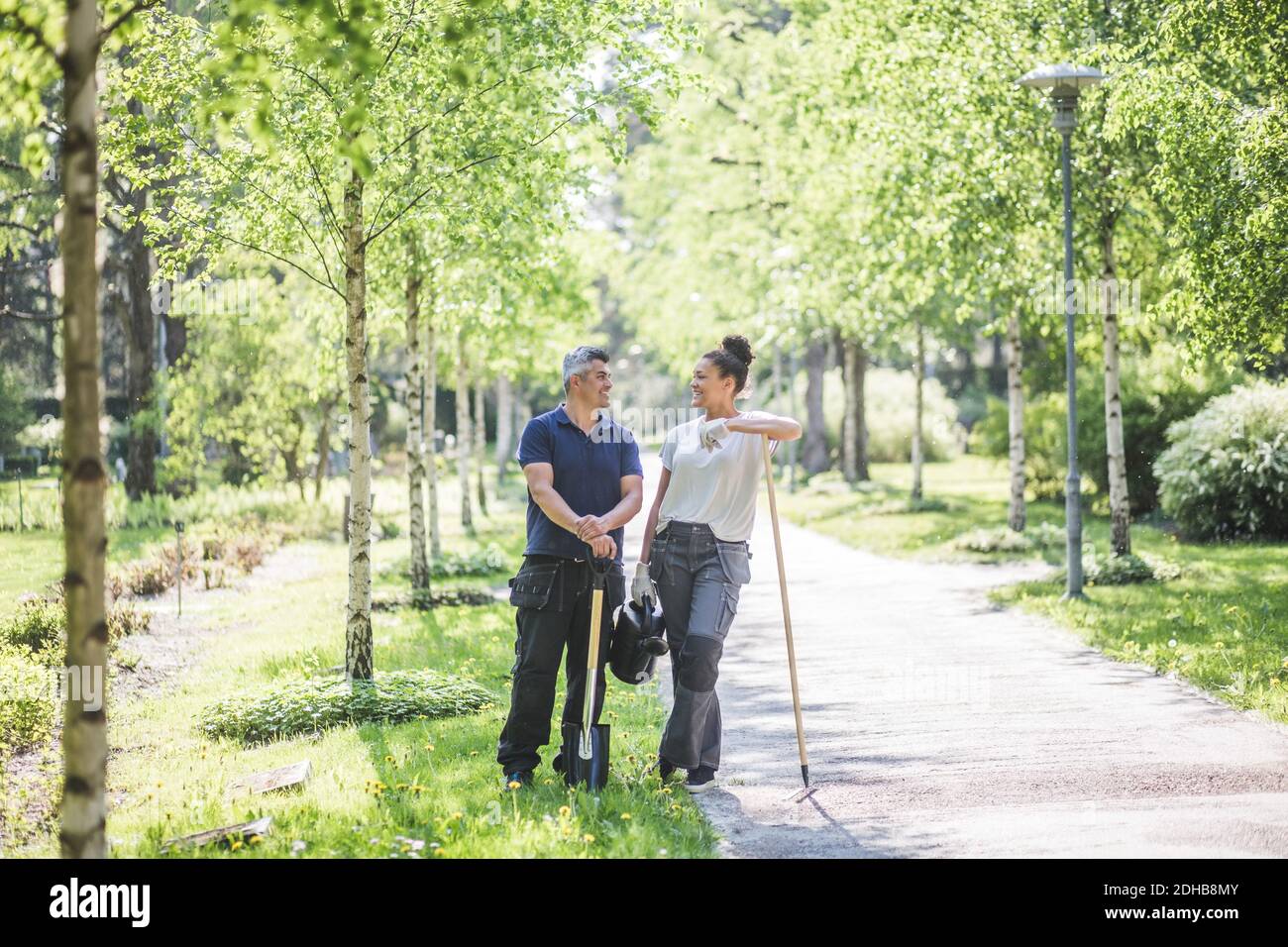 In voller Länge lächelnde weibliche Trainee mit Blick auf männlichen Lehrer Halten Gartengeräte auf Fußweg Stockfoto