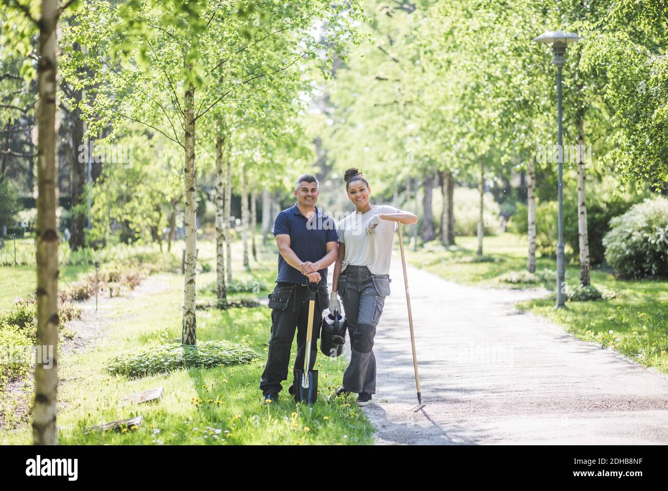Ganzkörperportrait mit lächelndem weiblichen Auszubildenden, der mit einem Mann steht Lehrer hält Gartengeräte auf Fußweg bei Stockfoto