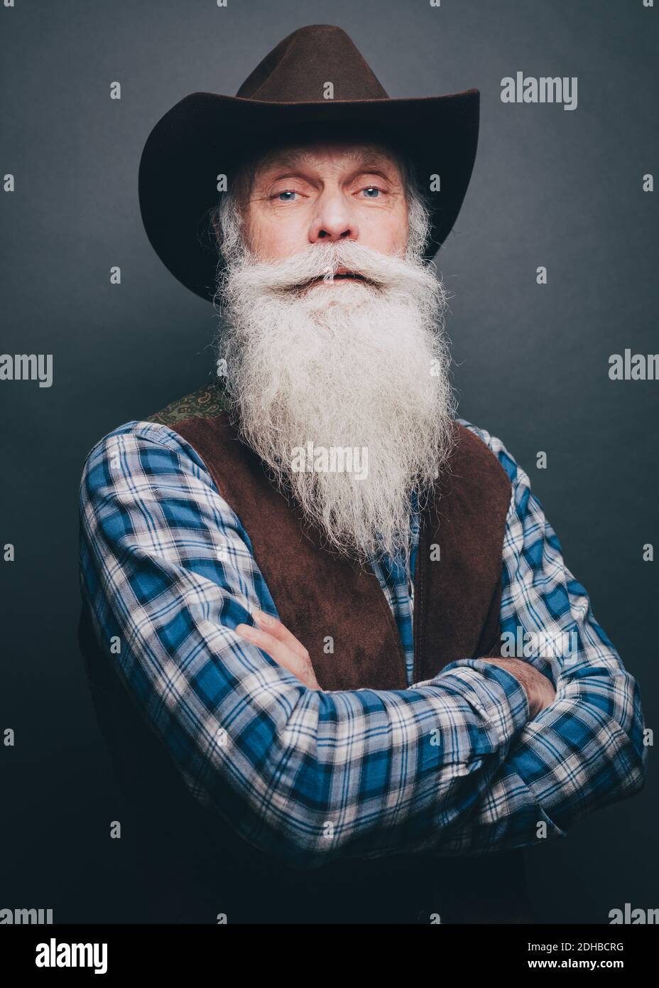 Bärtiger älterer Mann mit Cowboyhut und gekreuzten Armen Grauer Hintergrund Stockfoto