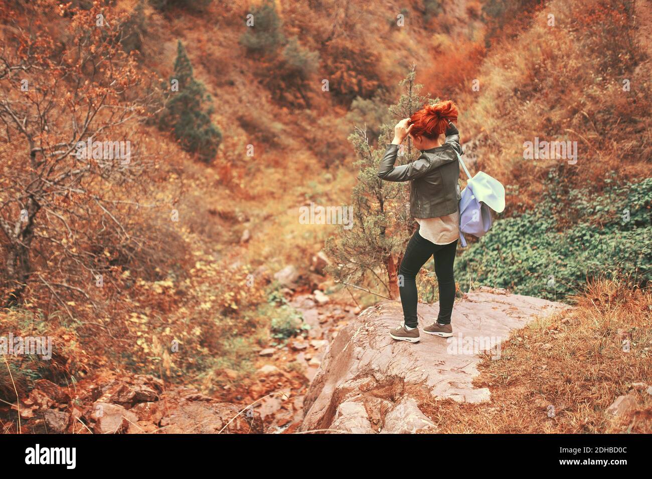 Weibliche Touristin im Herbst Bergwald. Rothaarige Frau, die ihr Haar auf dem Rand einer Klippe stehend tut. Herbsturlaub in die Natur. Blauer Wanderrucksack. Stockfoto