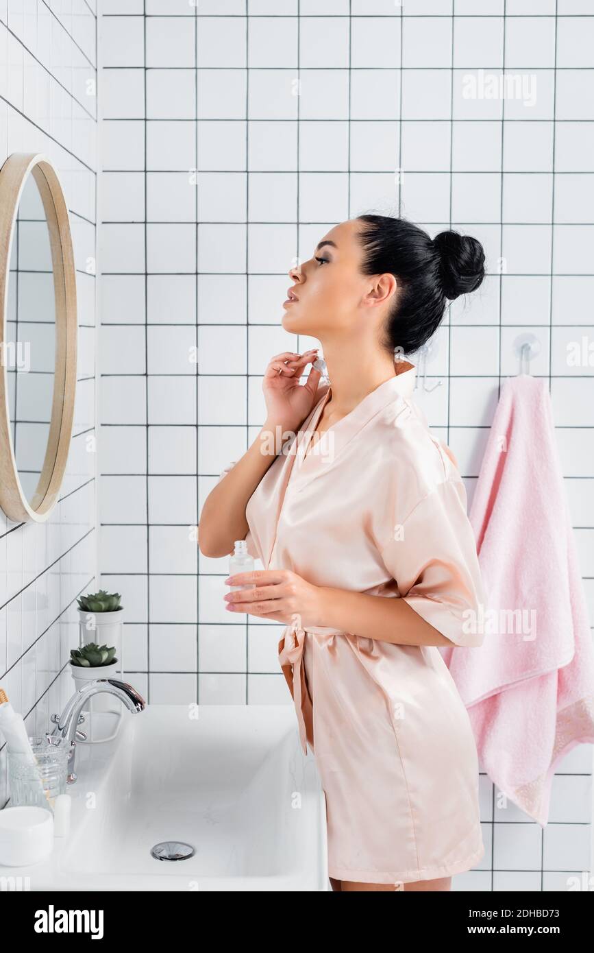 Junge Frau in Seide Bademantel Anwendung kosmetischen Serum in der Nähe des Spiegels Im Badezimmer Stockfoto