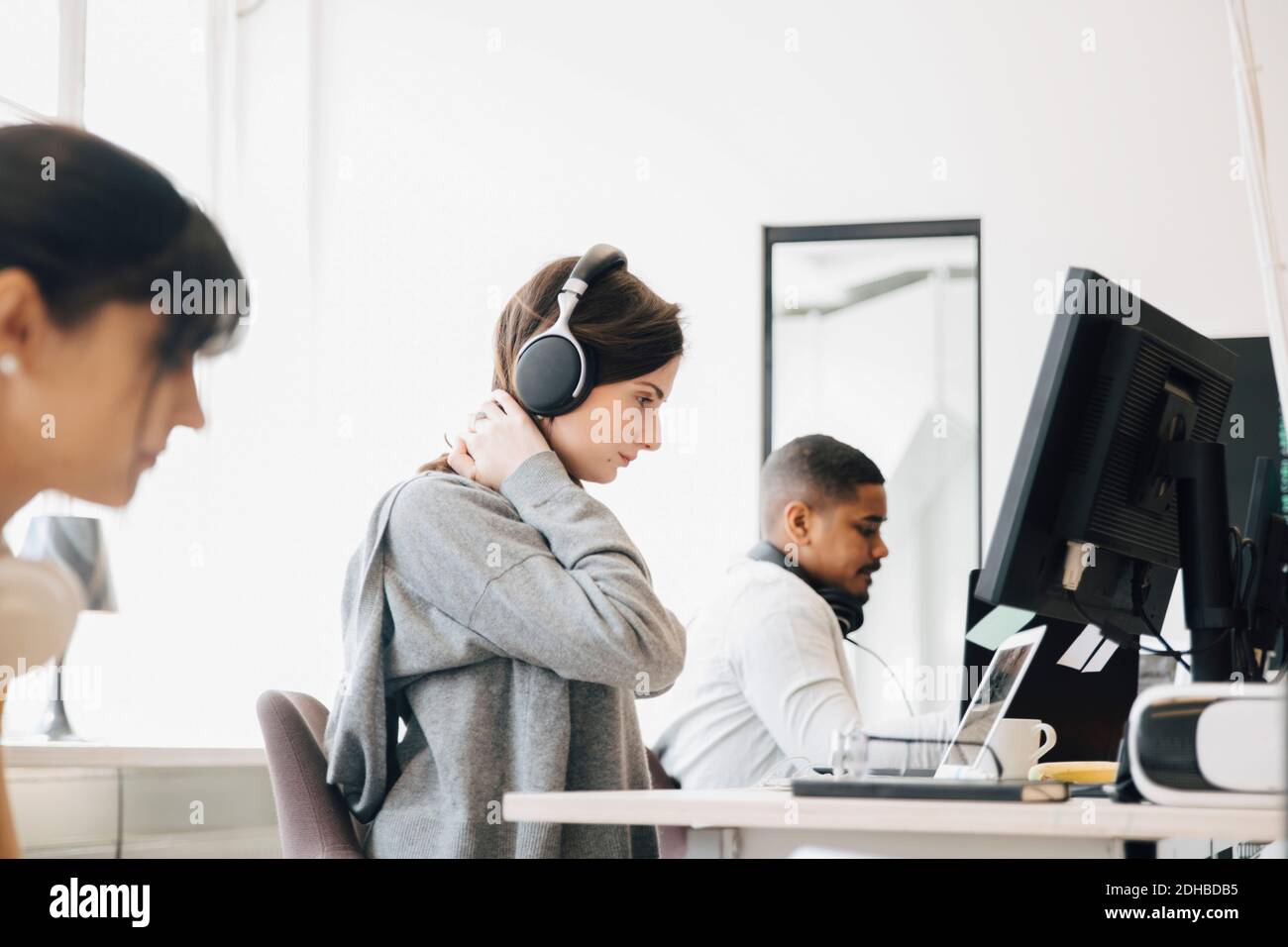 Fokussierte weibliche Programmierer mit Kopfhörer mit Laptop auf dem Schreibtisch während Sitzen mit Kollegen im Büro Stockfoto