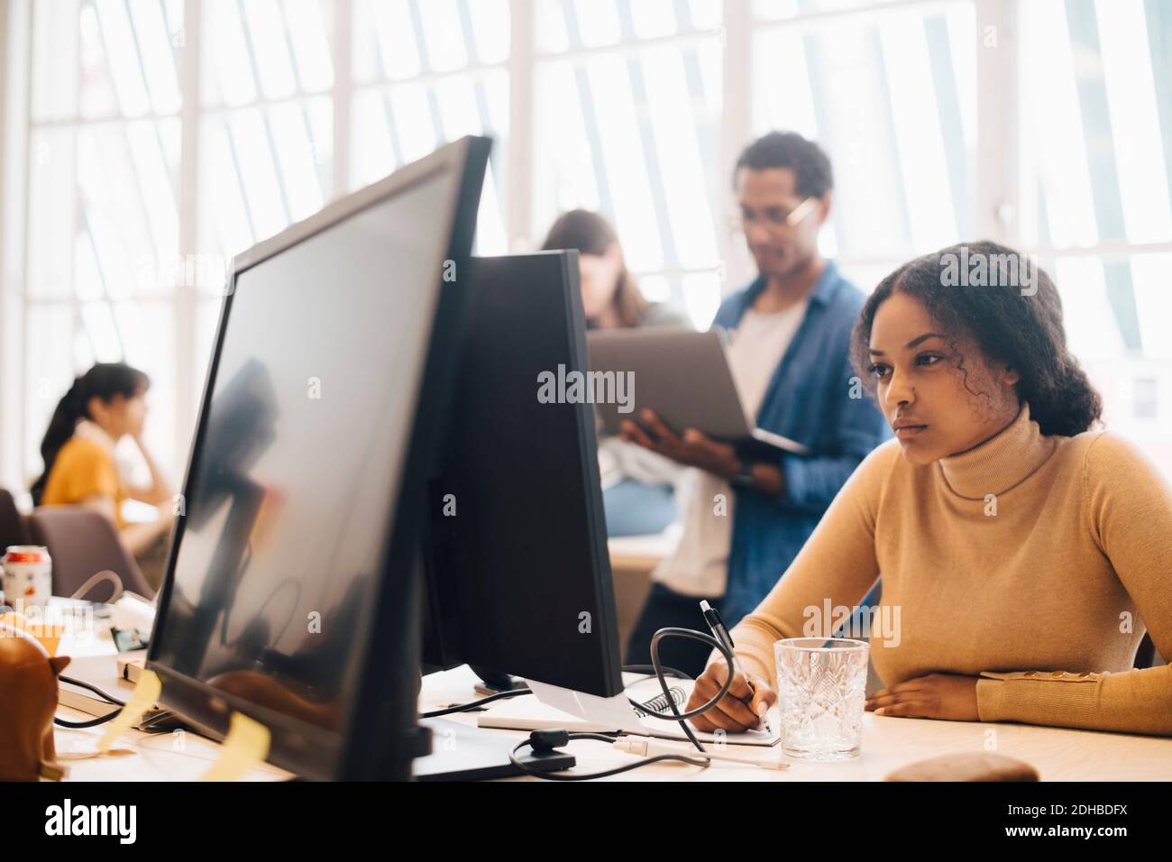 Fokussierte Geschäftsfrau mit Computer, während Mitarbeiter im Hintergrund arbeiten Büro Stockfoto
