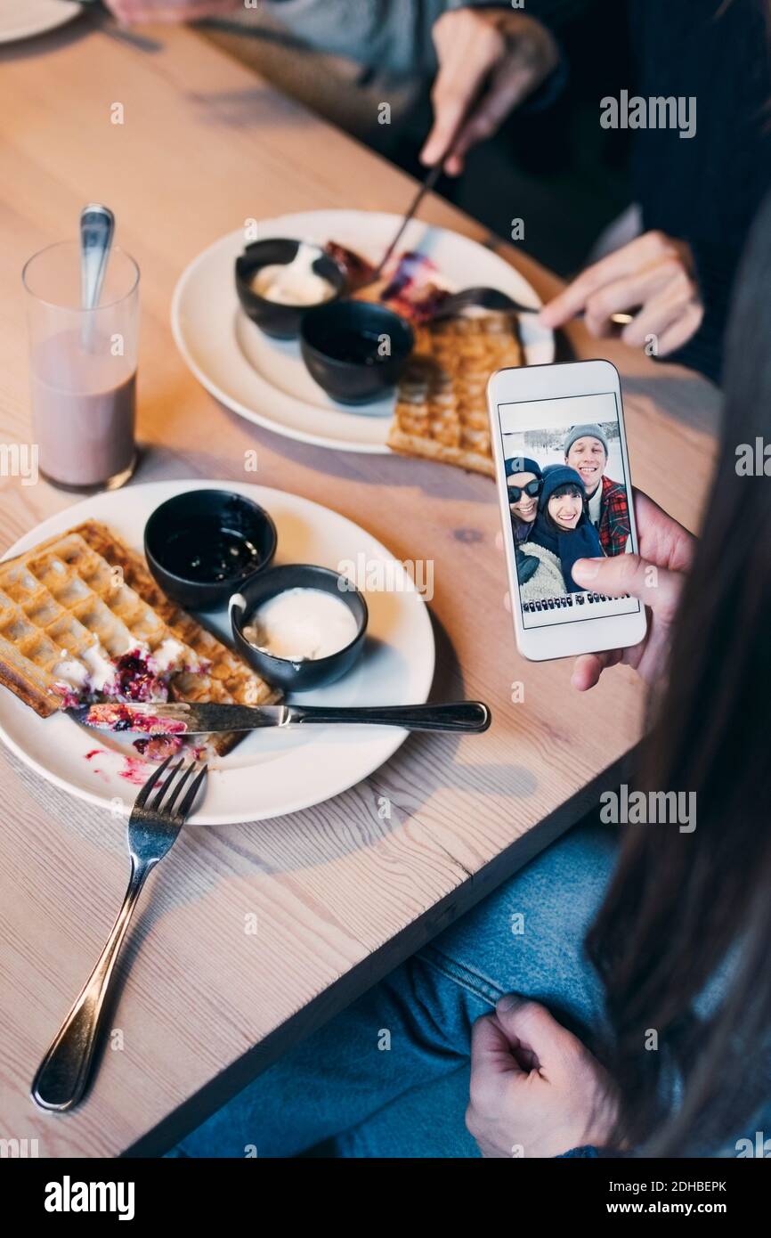Zugeschnittenes Bild einer Frau, die auf einem Smartphone auf ein Foto schaut Bildschirm beim Frühstück mit Freunden Stockfoto