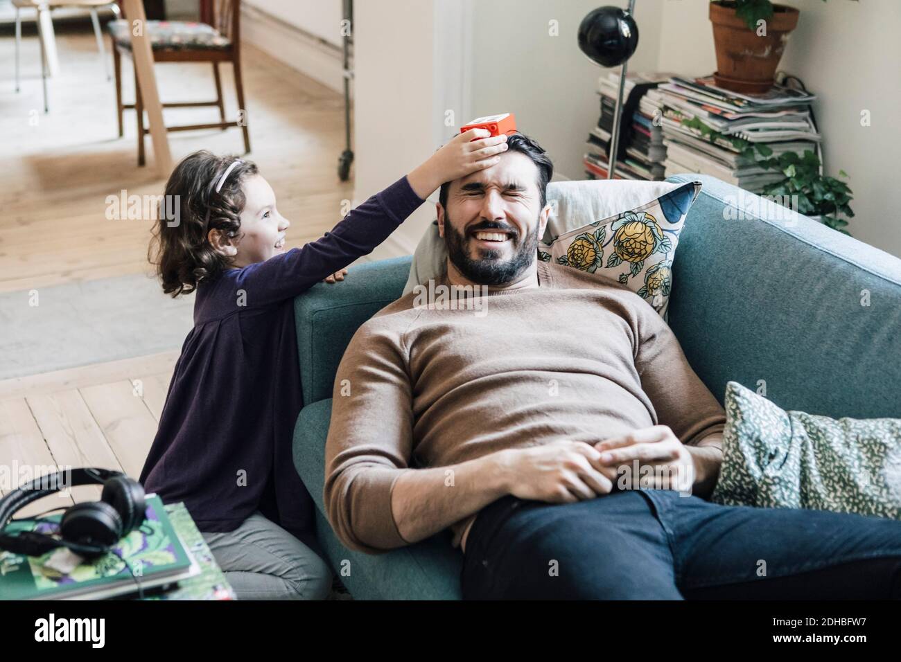 High-Angle-Ansicht von Mädchen halten Spielzeug Block auf Vater Kopf liegt auf dem Sofa zu Hause Stockfoto