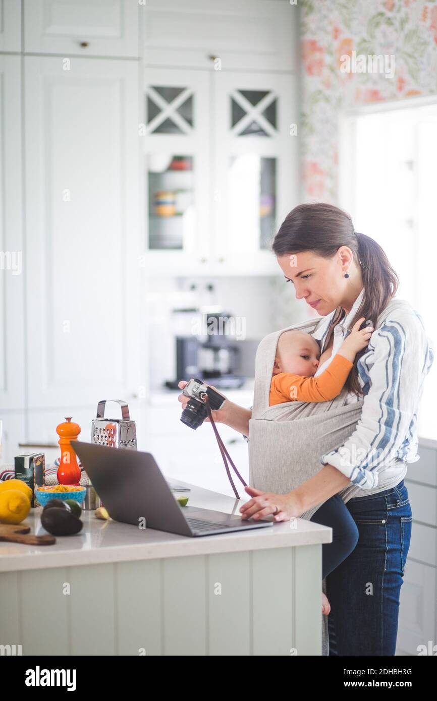 Seitenansicht der Lebensmittel-Blogger mit Kamera mit Laptop während Tragen Baby Mädchen in der Küche Stockfoto