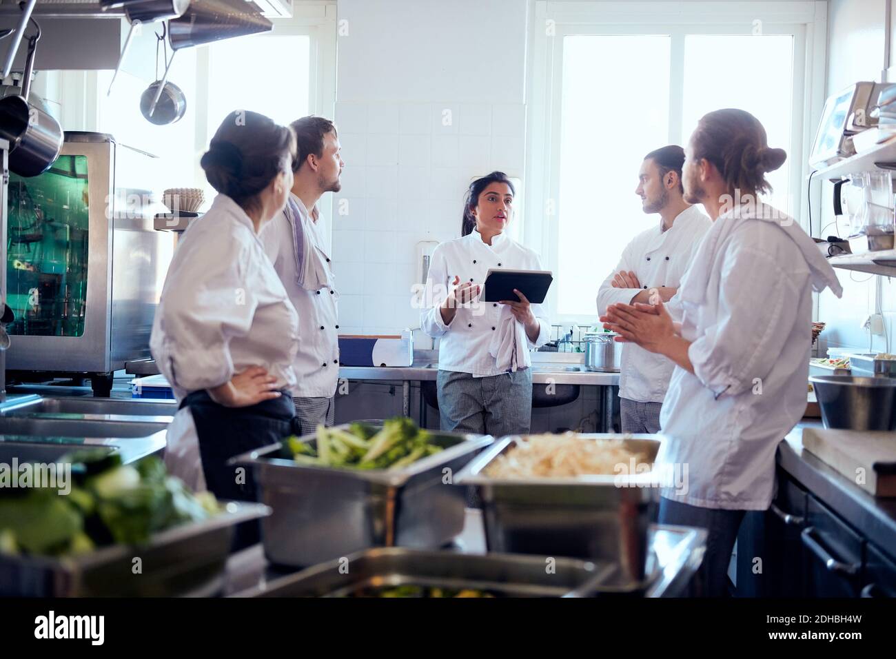 Köchin mit mittlerem Erwachsenenalter hält ein digitales Tablet und diskutiert mit Team in der Küche Stockfoto