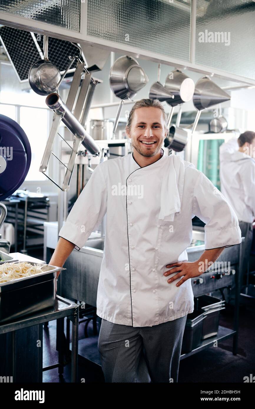 Porträt eines lächelnden männlichen Küchenchefs, der mit der Hand auf der Hüfte steht In der Küche Stockfoto