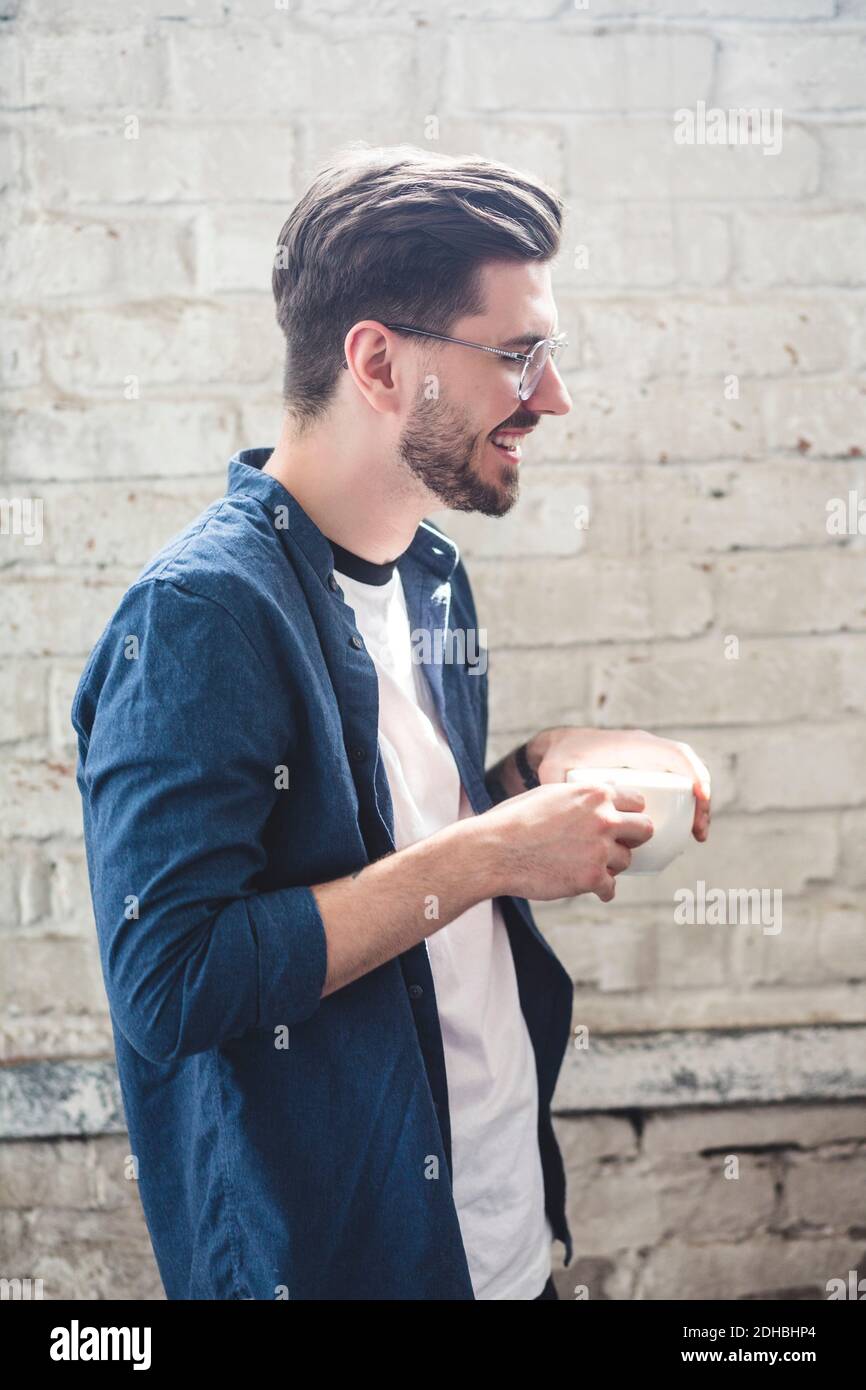 Seitenansicht des bärtig lächelnden Computerprogrammierers, der Kaffeetasse hält Im Büro stehen Stockfoto
