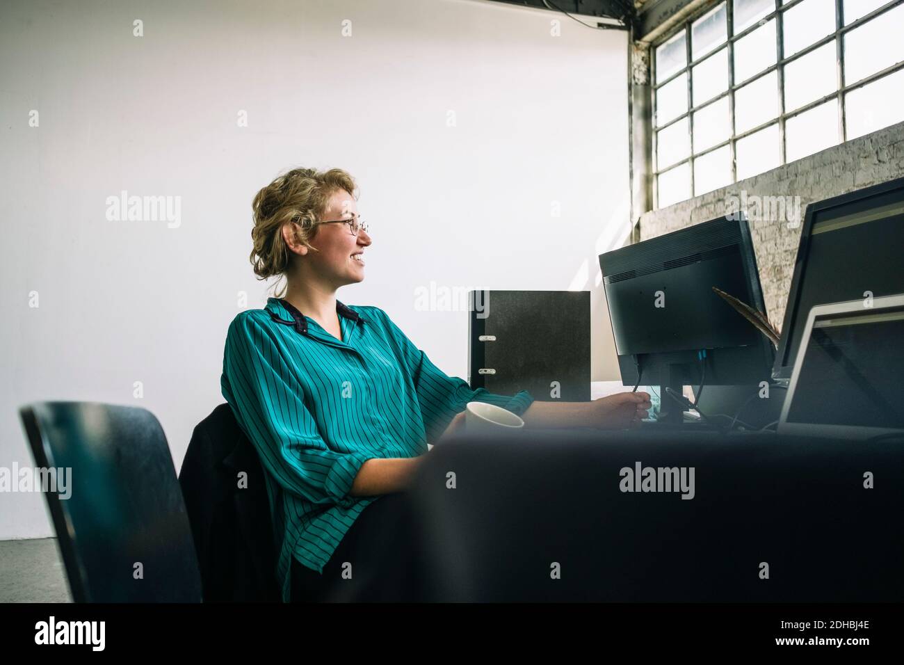 Nachdenkliche junge Geschäftsfrau lächelt und schaut weg, während sie ansitzt Schreibtisch im Kreativbüro Stockfoto
