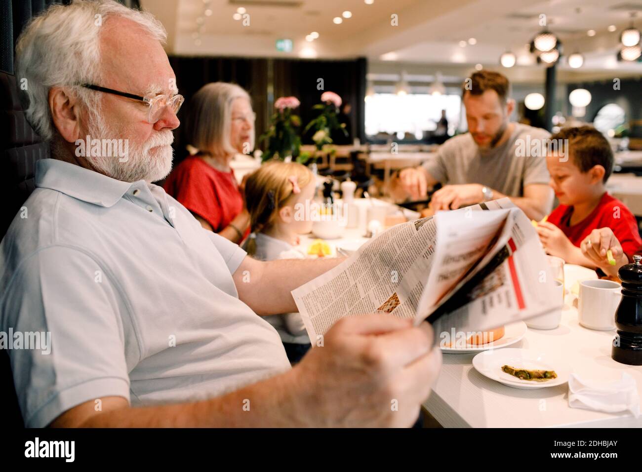 Älterer Mann liest Zeitung, während er Familie im Restaurant sitzt Frühstück Stockfoto