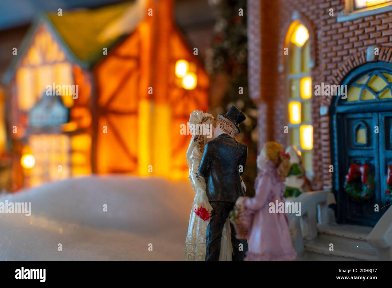 Eine Miniatur Weihnachtsdorf Ferienszene mit einer Braut und Bräutigam küssen vor einer Kirche, wie ein junges Blumenmädchen Uhren. Stockfoto