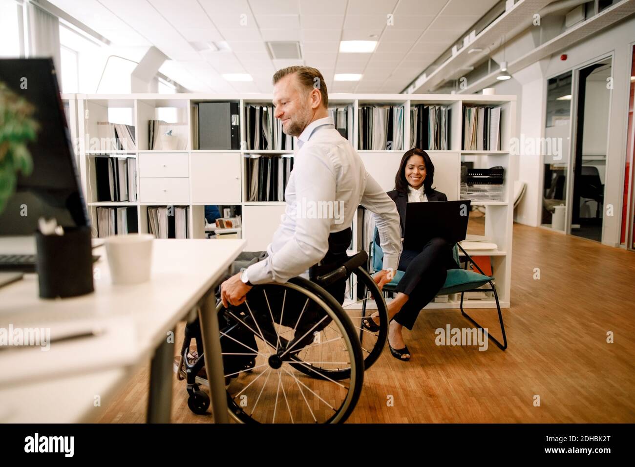 Behinderten männlichen professionellen sitzen im Rollstuhl, während lächelnd Geschäftsfrau mit Laptop am Arbeitsplatz Stockfoto