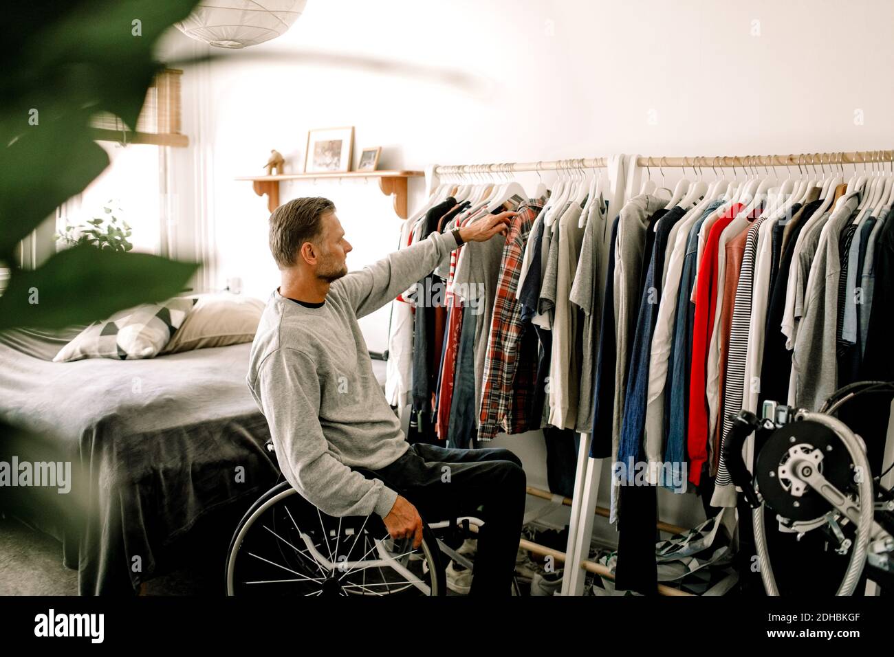 Reifer Mann sitzt auf Rollstuhl, während die Wahl Kleidung aus Rack Zu Hause Stockfoto