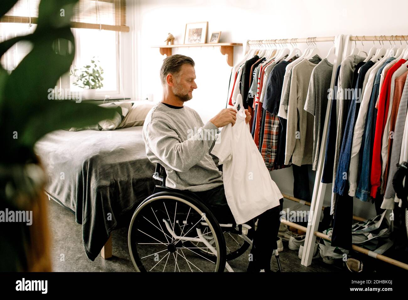 Seitenansicht des behinderten reifen Mannes mit weißem T-Shirt während Sitzen im Rollstuhl von Kleiderständer zu Hause Stockfoto