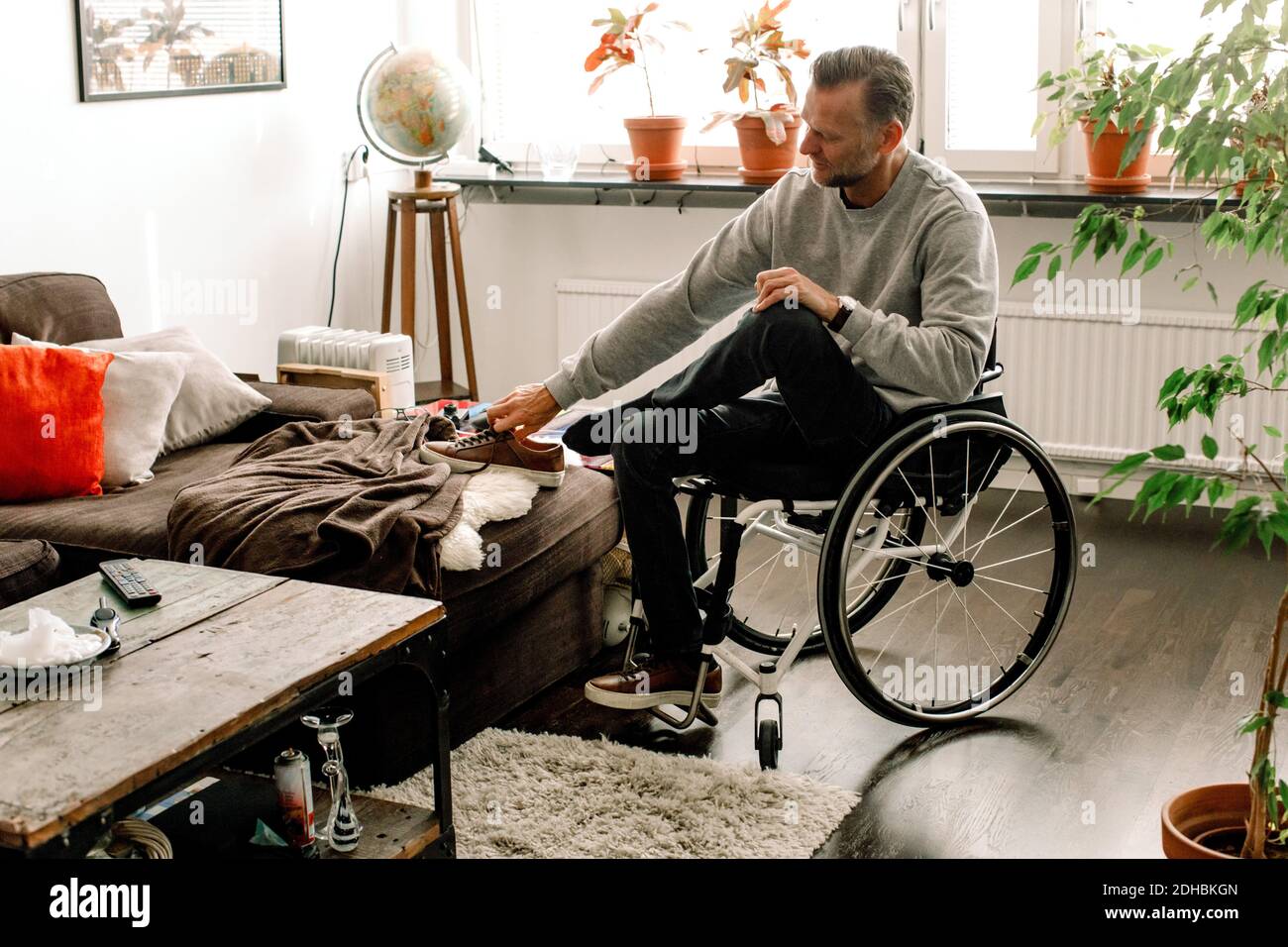 Volle Länge der behinderten reifen Mann trägt Schuh während des Sitzens Auf Rollstuhl im Wohnzimmer zu Hause Stockfoto