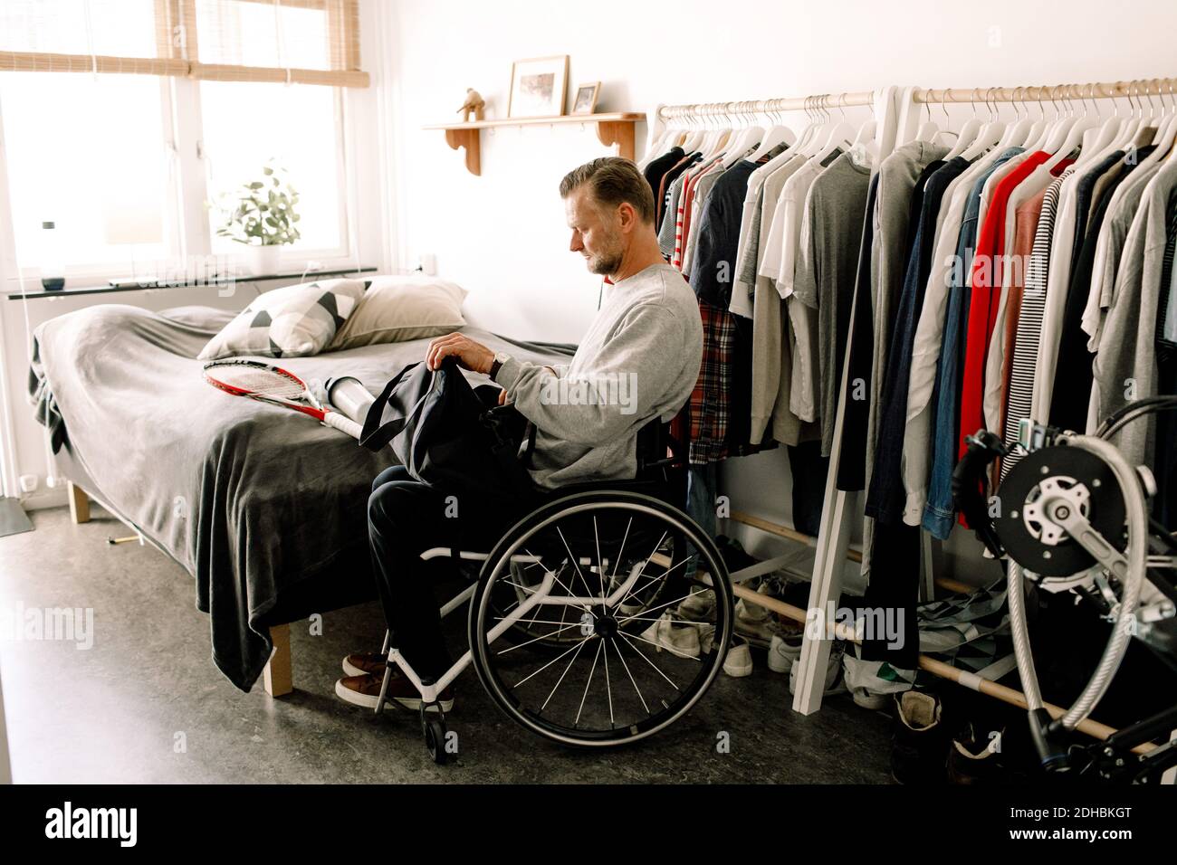 Seitenansicht des behinderten erwachsenen Mannes beim Einpacken des Tennisschlägers Tasche, während Sie zu Hause auf dem Rollstuhl sitzen Stockfoto