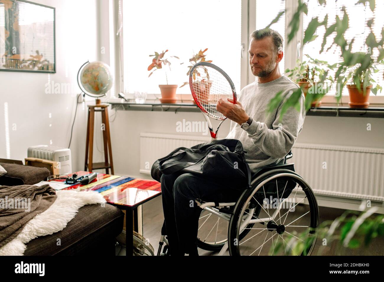 Nachdenklicher reifer Mann hält Tennisschläger, während er auf dem Rollstuhl sitzt Zu Hause Stockfoto
