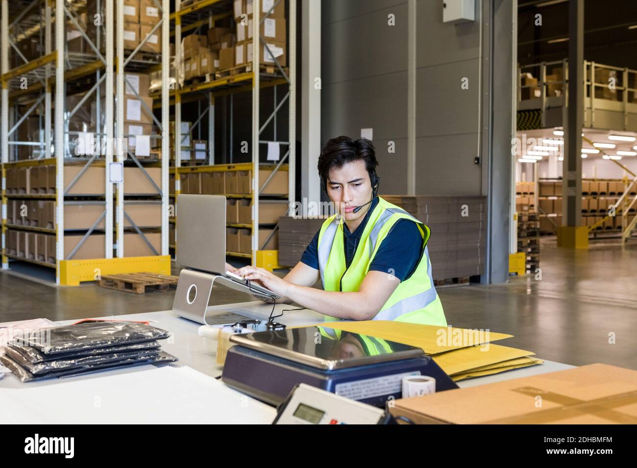 Selbstbewusster junger männlicher Kundendienstmitarbeiter mit Laptop während des Sitzens Am Schreibtisch im Lager Stockfoto Selbstbewusster junger männlicher Kundendienstmitarbeiter mit Laptop während des Sitzens Am Schreibtisch im Lager Stockfoto