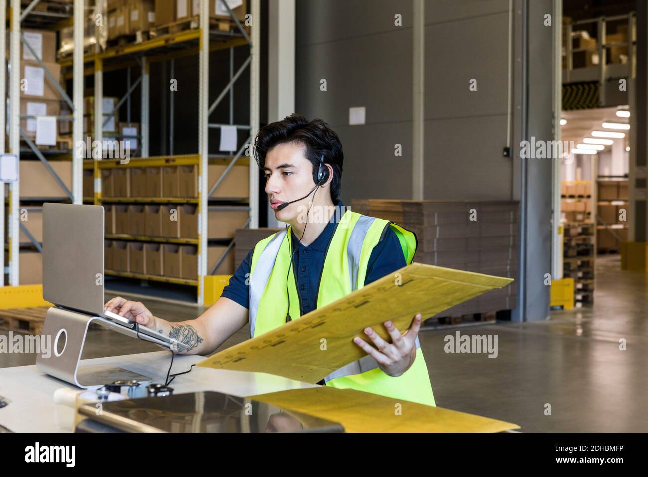 Selbstbewusster junger männlicher Kundendienstmitarbeiter mit Laptop während des Sitzens Mit Paket am Schreibtisch im Lager Stockfoto Selbstbewusster junger männlicher Kundendienstmitarbeiter mit Laptop während des Sitzens Mit Paket am Schreibtisch im Lager Stockfoto