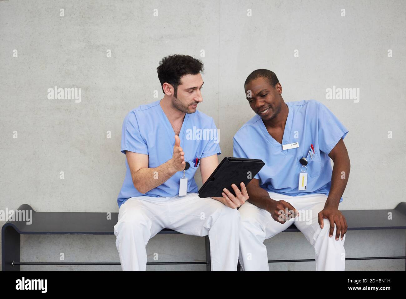 Männliche Krankenschwestern diskutieren über digitale Tablette, während sie gegen grau sitzen Wand im Flur im Krankenhaus Stockfoto