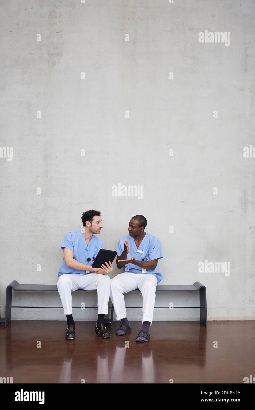 Volle Länge der männlichen Krankenschwestern diskutieren über digitale Tablette während Sitzen gegen graue Wand im Flur im Krankenhaus Stockfoto