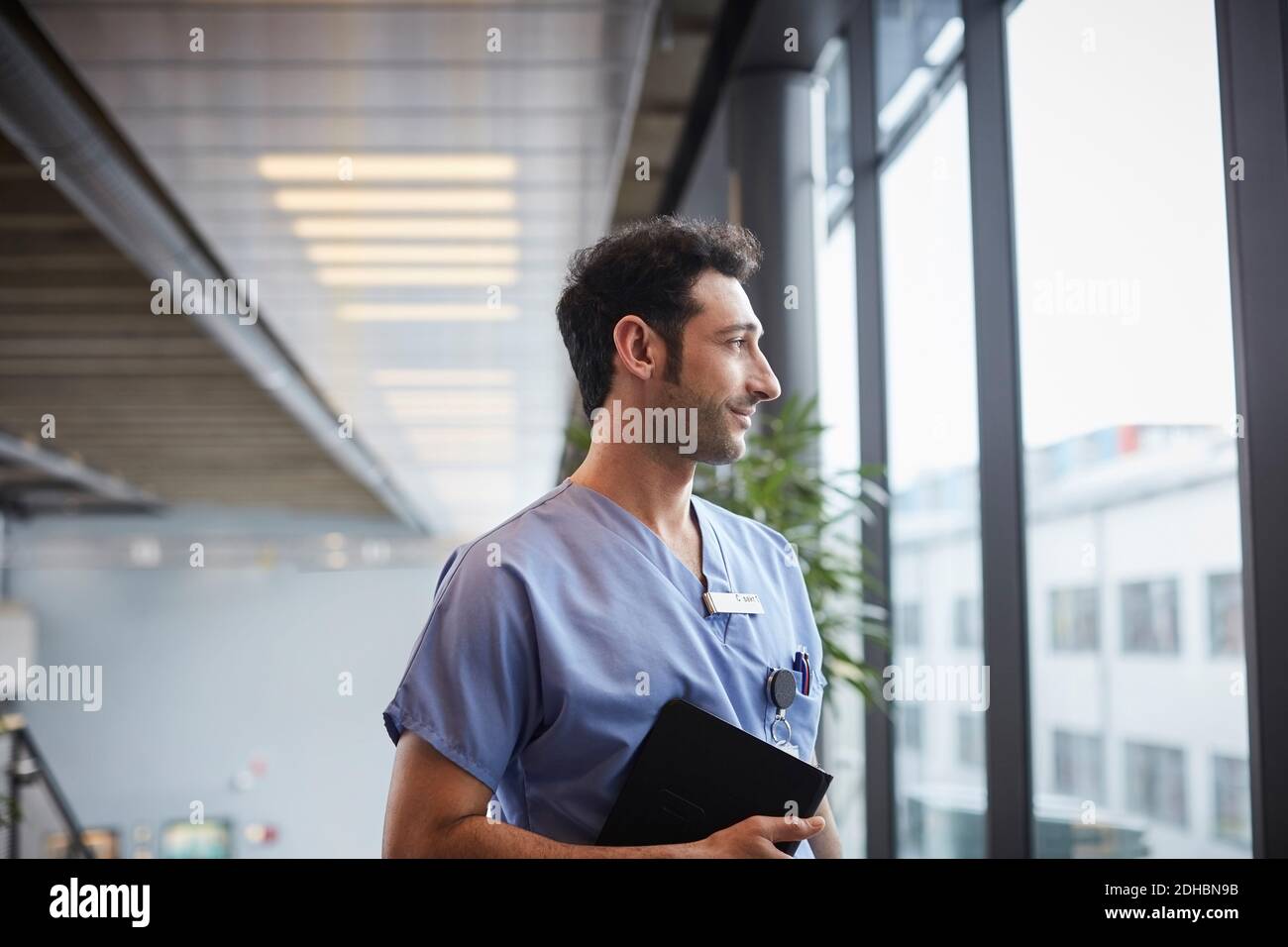 Nachdenkliche junge Krankenschwester hält digitale Tablette beim Blick durch Fenster im Krankenhaus Stockfoto
