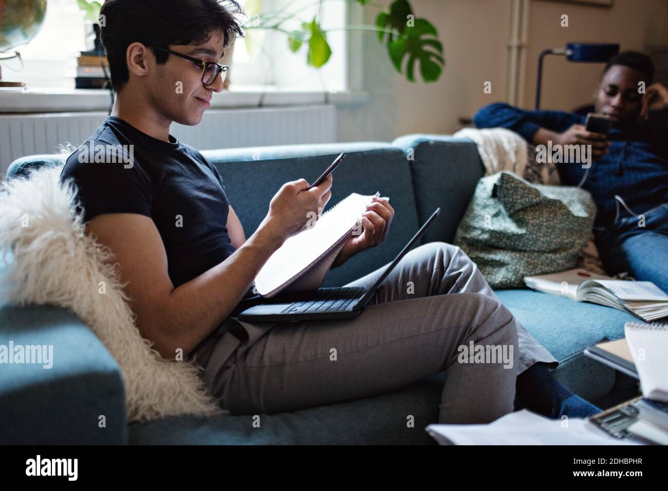 Junger Mann, der studiert, während Freund soziale Medien auf dem Sofa nutzt Zu Hause Stockfoto